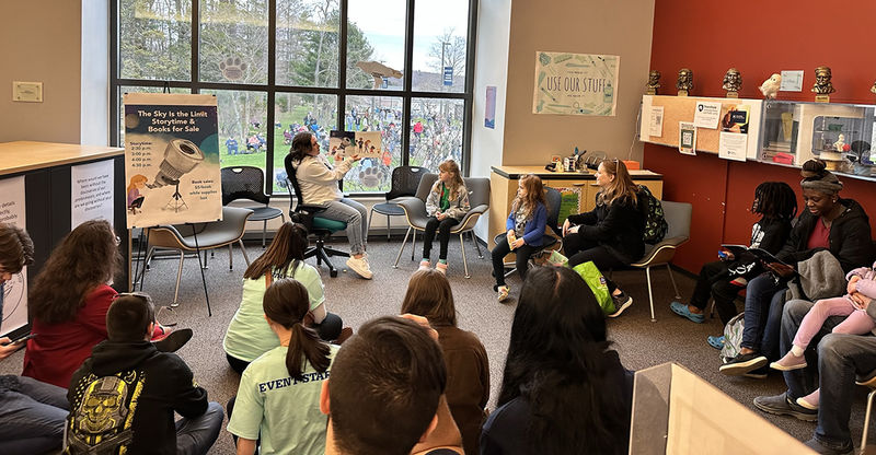 A woman holding up a book while reading a story to a room of people.
