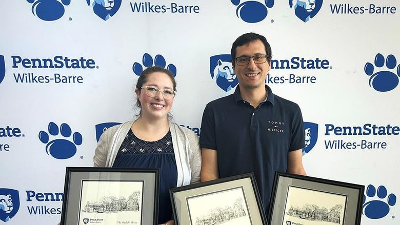 A woman and a man standing against a backdrop with Penn State Wilkes-Barre's logo and holding framed awards.