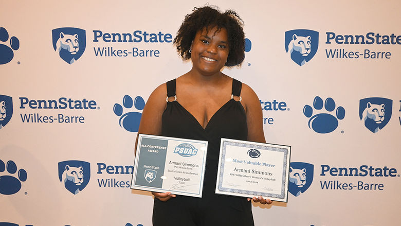 A woman holding two certificates standing against a backdrop with the Penn State Wilkes-Barre logo.