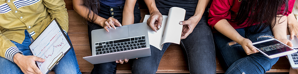 Four individuals sitting side by side on a bench; close up of their laps with clipboard, laptop computer, pad, and tablet.