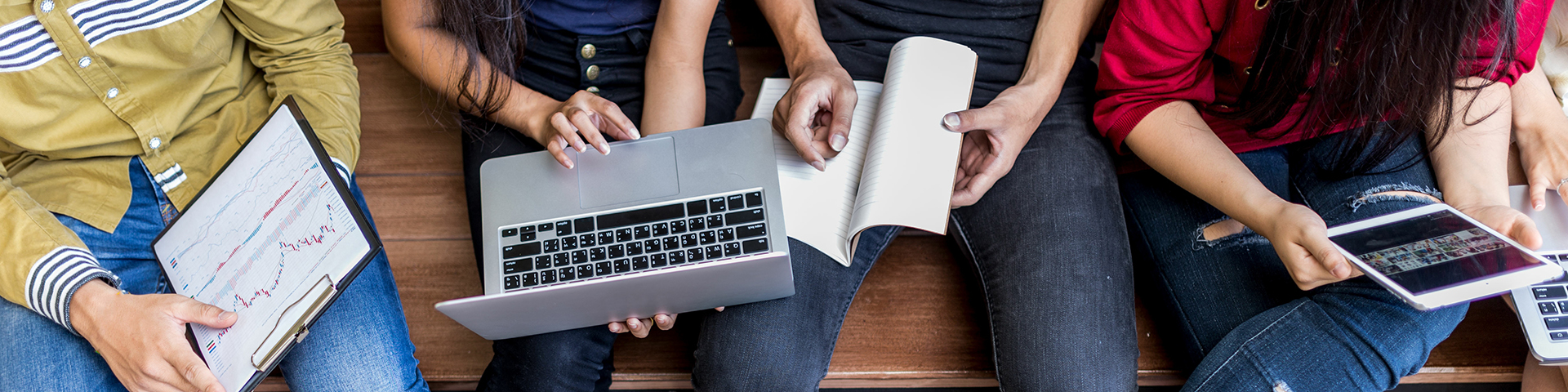 Four individuals sitting side by side on a bench; close up of their laps with clipboard, laptop computer, pad, and tablet.