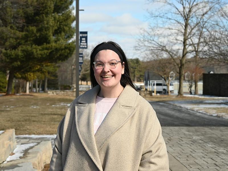 A smiling woman wearing glasses and a headband standing outside on a walkway, with Penn State Wilkes-Barre flags lining the walkway behind her