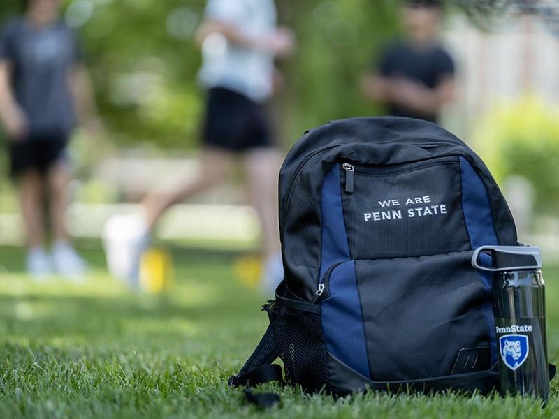 A backpack with the words "We Are Penn State" next to a water bottle with the Penn State logo sitting on a grassy field.