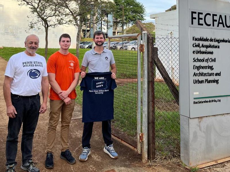 Three people, one holding a Penn State Wilkes-Barre shirt, stand outside a building in Brazil.
