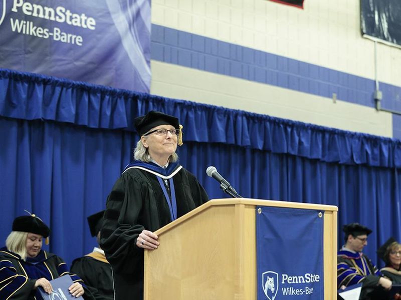 A woman in academic regalia speaks at a podium bearing the Penn State Wilkes-Barre logo.