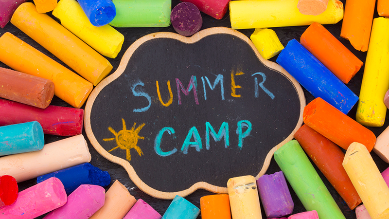 A pile of brightly colored chalk, with a decorative chalkboard resting on top with the words "Summer Camp"