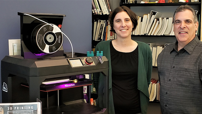 Jonathan Pineno (Friedman Art Gallery director) and Megan Mac Gregor (librarian) standing in front of the Nesbitt Library's 3D printer; two of the small green widgets are sitting on top.