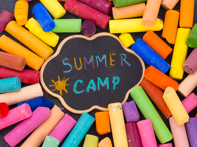 A pile of brightly colored chalk upon which is lying a small decorative slate chalkboard. The text on the chalkboard is "Summer Camp"