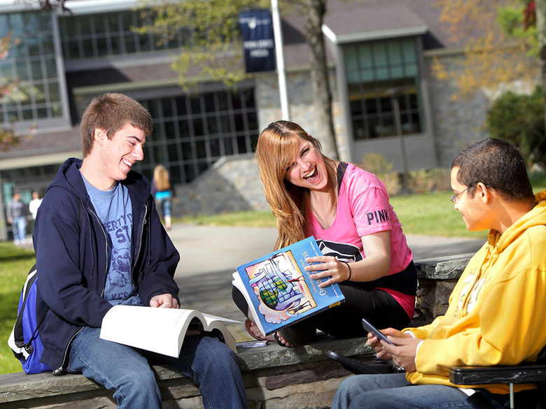 Three laughing students sitting on the wall in front of Hayfield House with the Academic Commons in the background