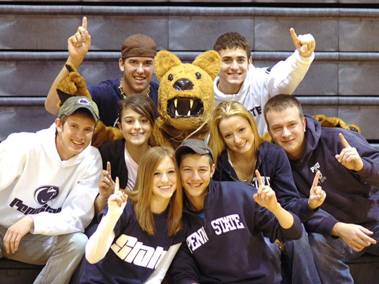 Students sitting on bleachers and gathered around the Nittany Lion mascot, facing forward and holding up their index fingers in the “we're number one” sign