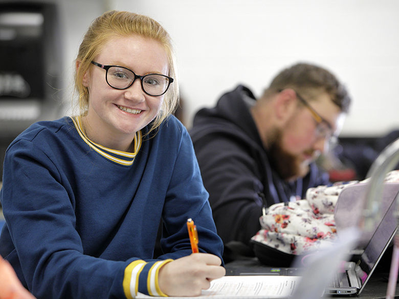In the classroom: a female student with red hair and glasses is looking at the camera, while a male student in the background is looking down at his work.