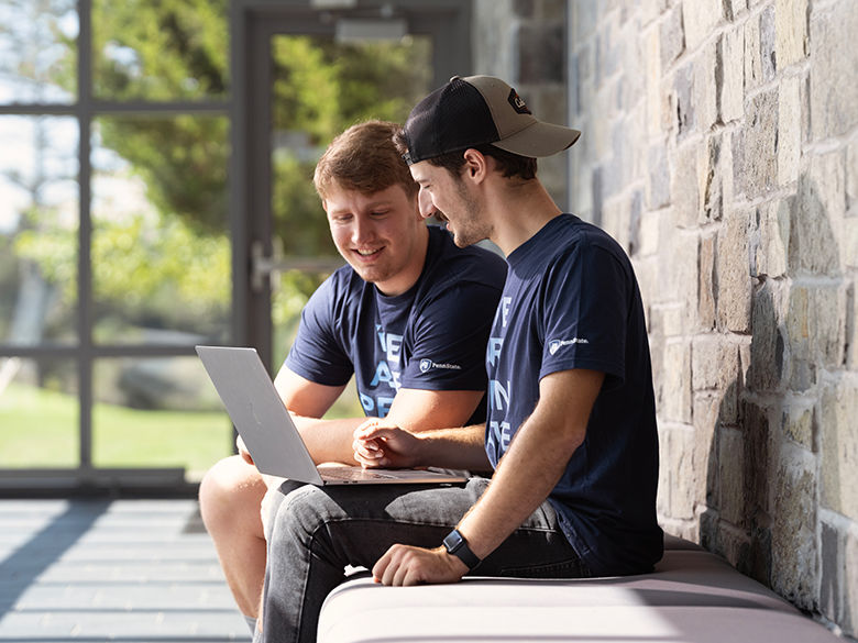 Two male students looking at a laptop computer while sitting on a sunlit bench in the foyer of the Academic Commons