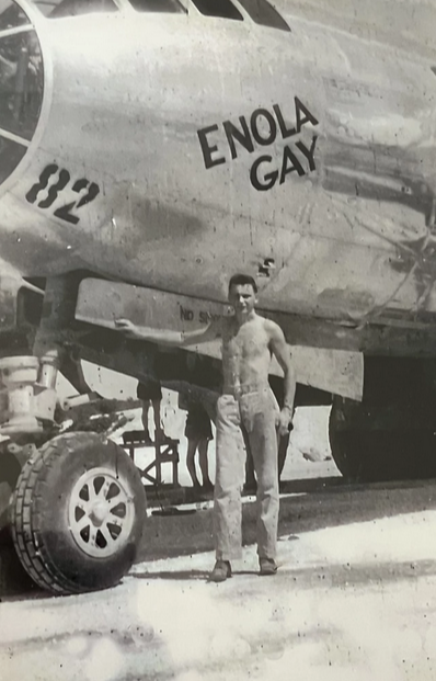 A black-and-white image of a man standing next to a plane with the words "Enola Gay" on its side