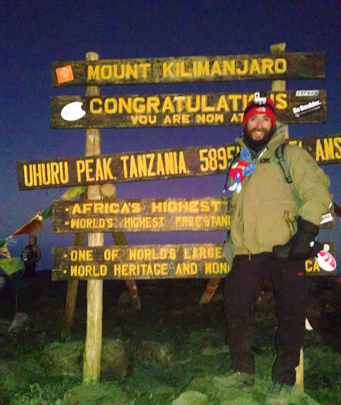 A man in winter gear standing in front of a wooden sign that says "Mount Kilimanjaro."