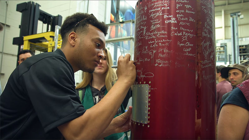 Participants signing the rocket in silver ink; the metallic red surface is covered in signatures.