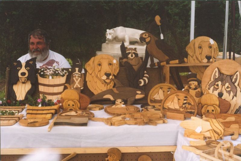 A man sits behind a table filled with wood carvings.