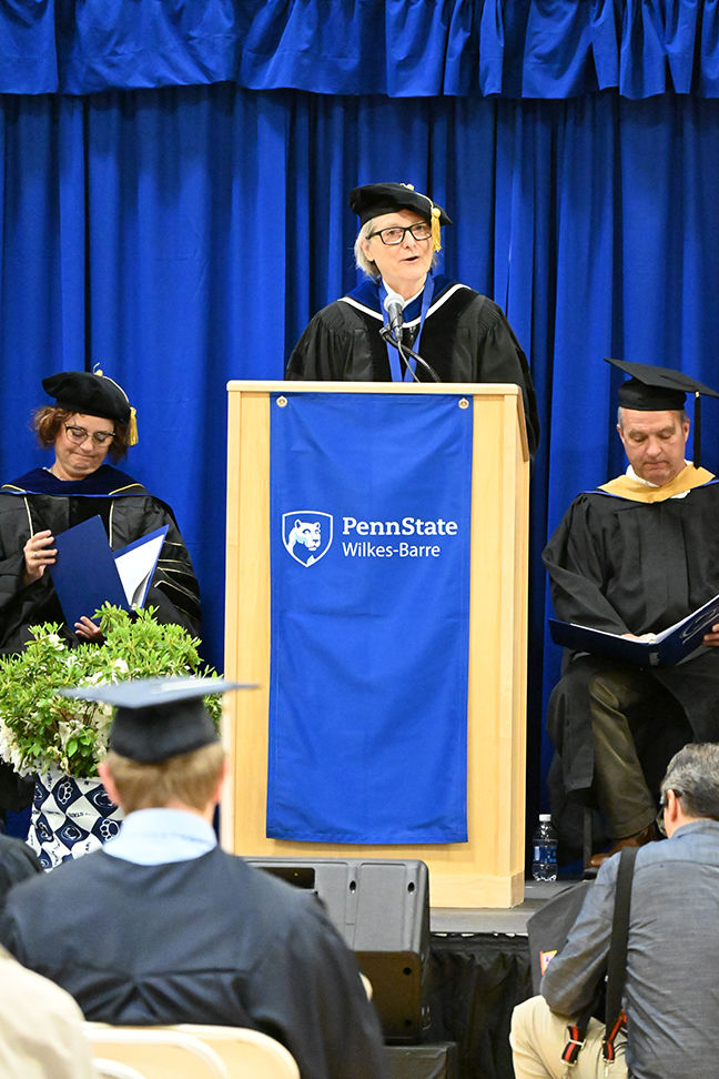 A woman in academic regalia speaks from the podium.
