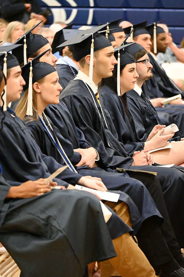 Graduates in caps and gowns listen to a speaker.