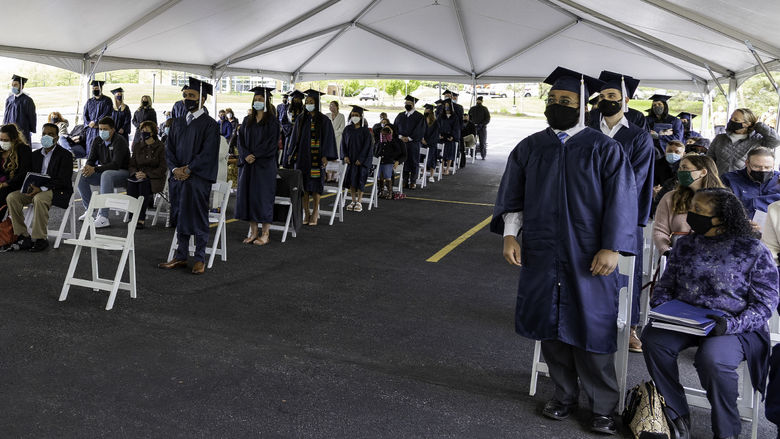 Students dressed in caps and gowns and wearing masks stand while audience members remain seated.