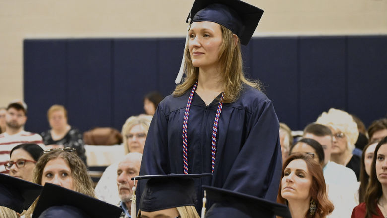 A woman wearing a cap and gown as well as a cord indicating her veteran status stands among seated graduates and audience members