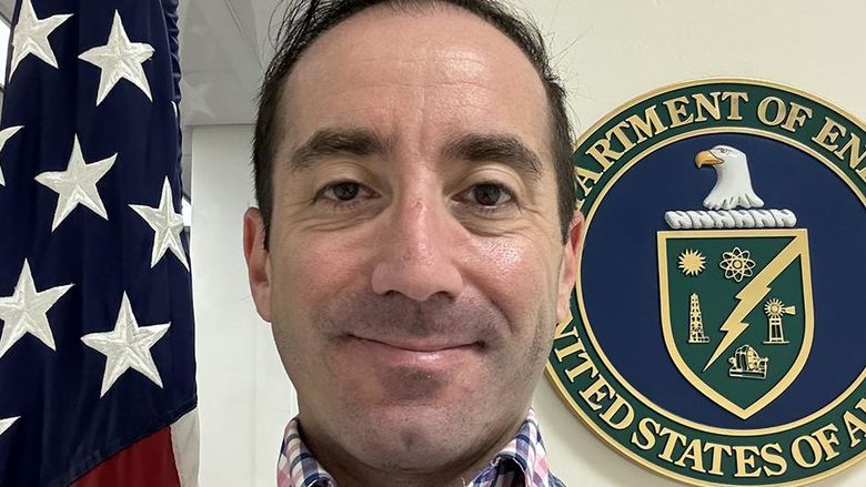 A man standing between a U.S. flag and a Department of Energy seal.