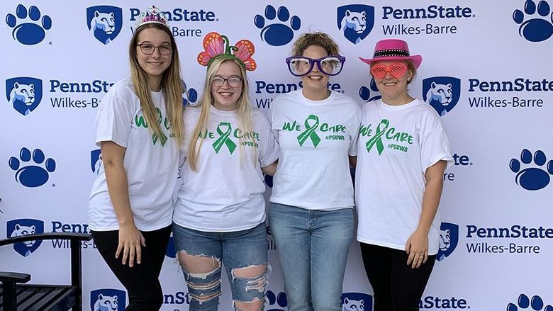 A group of four women, one in oversized glasses, stand against a backdrop with the Penn State Wilkes-Barre logo.