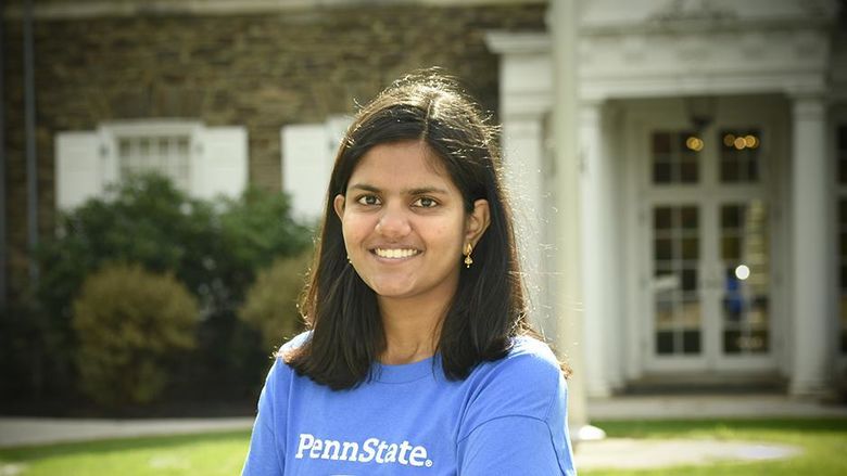 A student wearing a blue shirt with the Penn State mark, standing in front of a historic building.