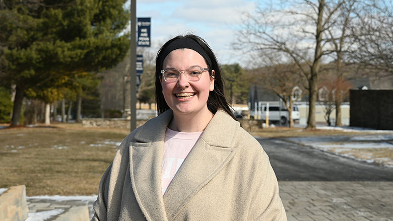 A smiling woman wearing glasses and a headband standing outside on a walkway, with Penn State Wilkes-Barre flags lining the walkway behind her