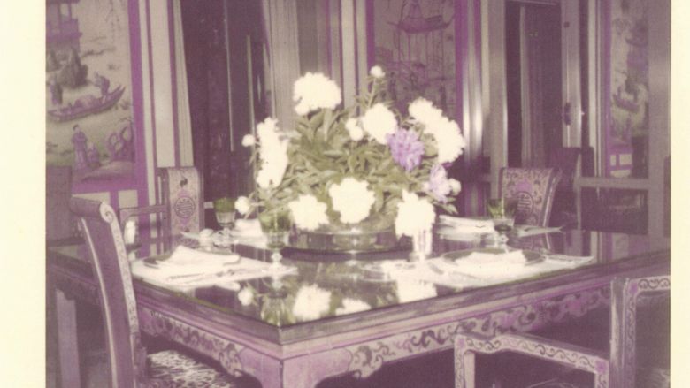 A historic photo of a room featuring a dining table and chairs with a large bouquet atop the table.