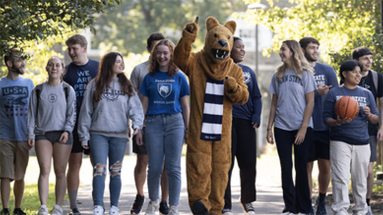A group of students strolling with the Nittany Lion mascot down a leafy pathway on the Penn State Wilkes-Barre campus