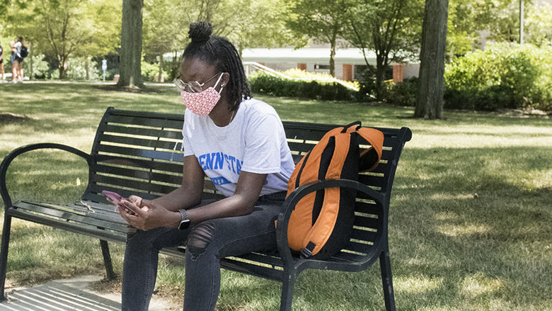 A Penn State student wearing a face mask looks at her phone while sitting on a campus bench.