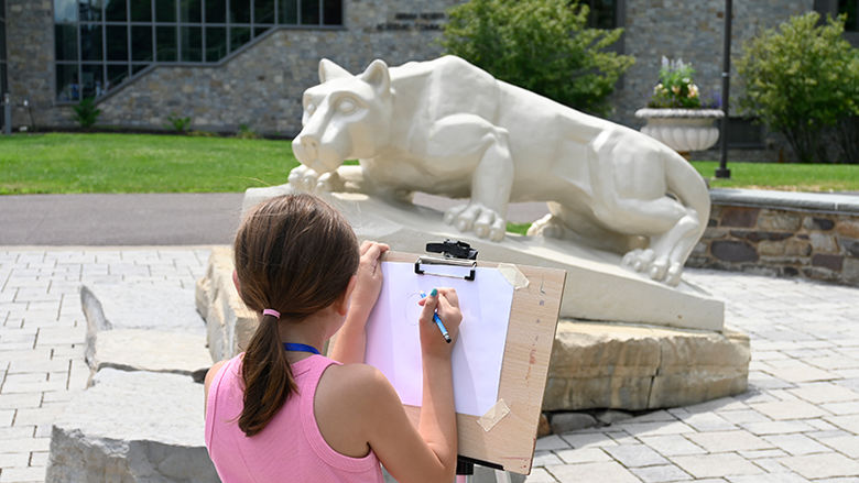A young girl with her back to the camera drawing on a sketch pad on an easel in front of the Nittany Lion shrine.