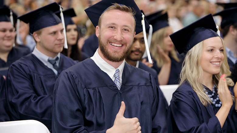 Grinning graduate in academic robe flashing the "thumbs up" sign at the camera