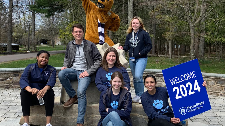 A diverse group of students sitting on the Lion Shrine with the Nittany Lion Mascot. One is holding a Welcome 2024 sign.
