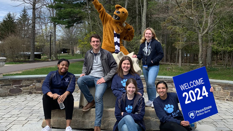 A group of students, along with the Nittany Lion mascot, sitting on the lion shrine. One student is holding a "Welcome 2024" sign.