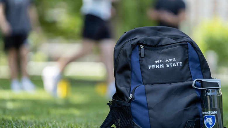 A backpack with the words "We Are Penn State" next to a water bottle with the Penn State logo sitting on a grassy field.
