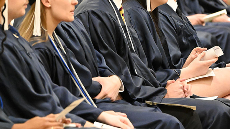 Graduates in caps and gowns listen to a speaker.