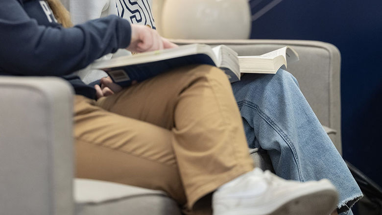 An image showing the legs of two students with textbooks on their laps, as one student gestures at a book.