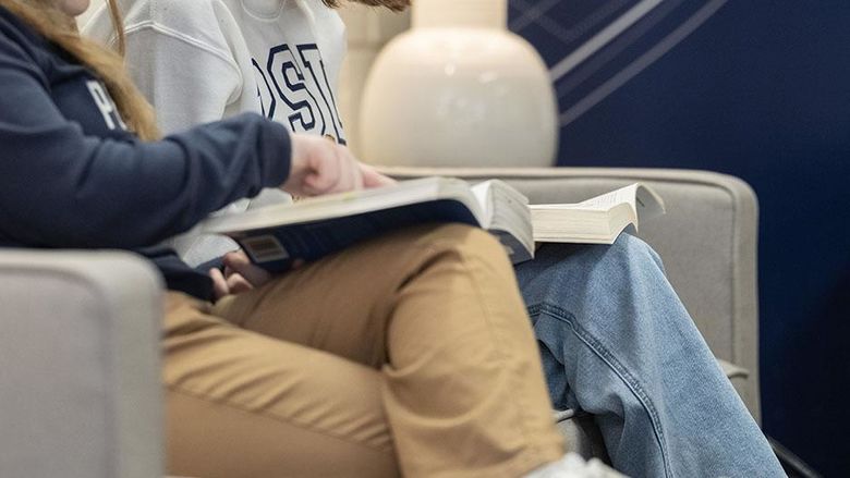 An image showing the legs of two students with textbooks on their laps, as one student gestures at a book.