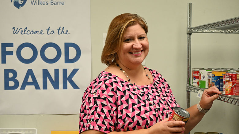 A woman standing next to a rack of canned goods, with the words "food bank" on a sign behind her.