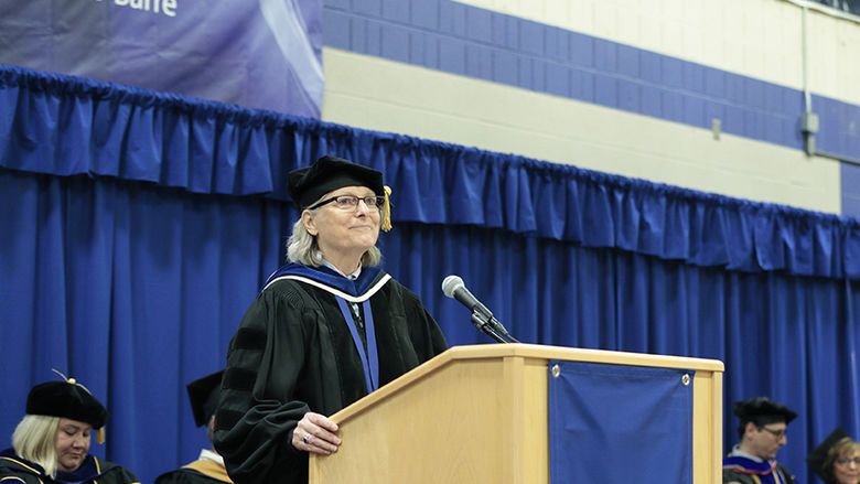 A woman in academic regalia speaks at a podium bearing the Penn State Wilkes-Barre logo.