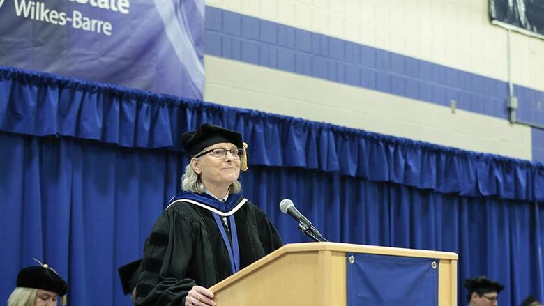 A woman in academic regalia speaks at a podium bearing the Penn State Wilkes-Barre logo.