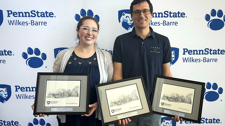 A woman and a man standing against a backdrop with Penn State Wilkes-Barre's logo and holding framed awards.