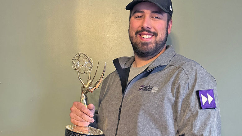 A man in a black baseball cap and gray jacket holding an Emmy statuette.