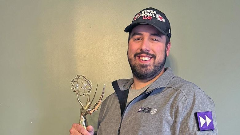 A man in a black baseball cap and gray jacket holding an Emmy statuette.