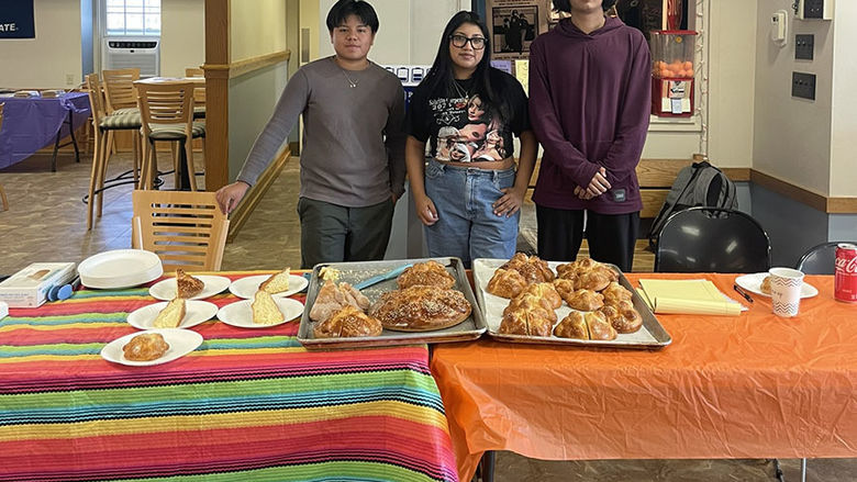 Three people standing behind a table with food placed on a colorful tablecloth.