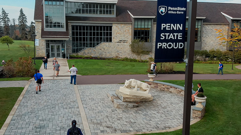 A view looking down at a campus building with large windows, a walkway with pavers and a Nittany Lion statue, with students walking throughout the photo.