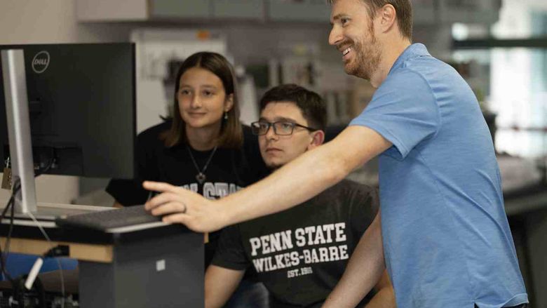 A man standing at right assists two seated students working on a computer.