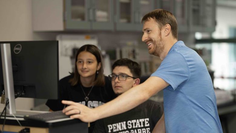 A man standing at right assists two seated students working on a computer.