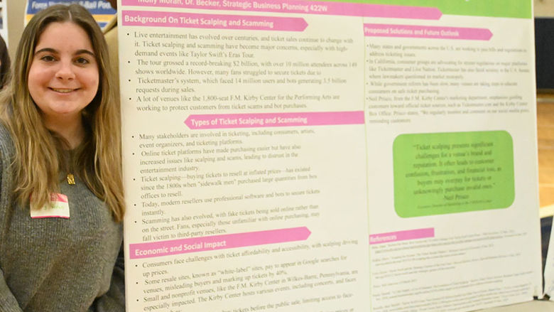 A woman stands at left next to a large research poster on a table.